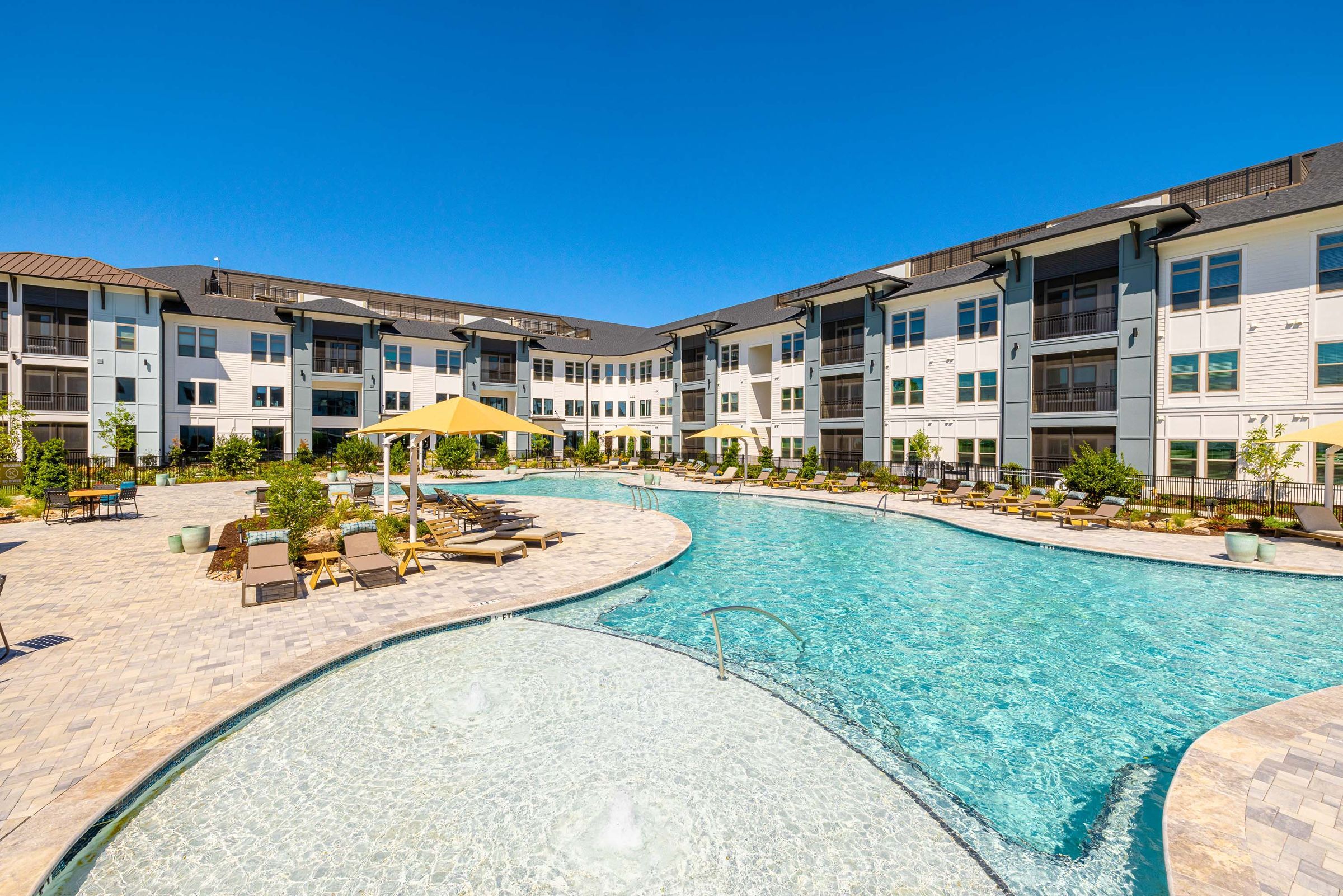 Botanic Luxury Apartments A modern apartment complex with a large outdoor pool, lounge chairs, and yellow umbrellas on a sunny day.