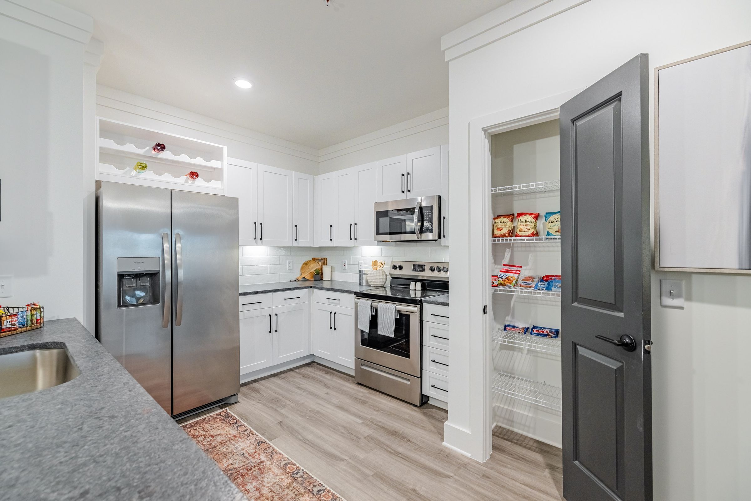 Botanic Luxury Apartments Modern kitchen with stainless steel appliances, white cabinetry, a light gray countertop, and an open pantry with snacks.